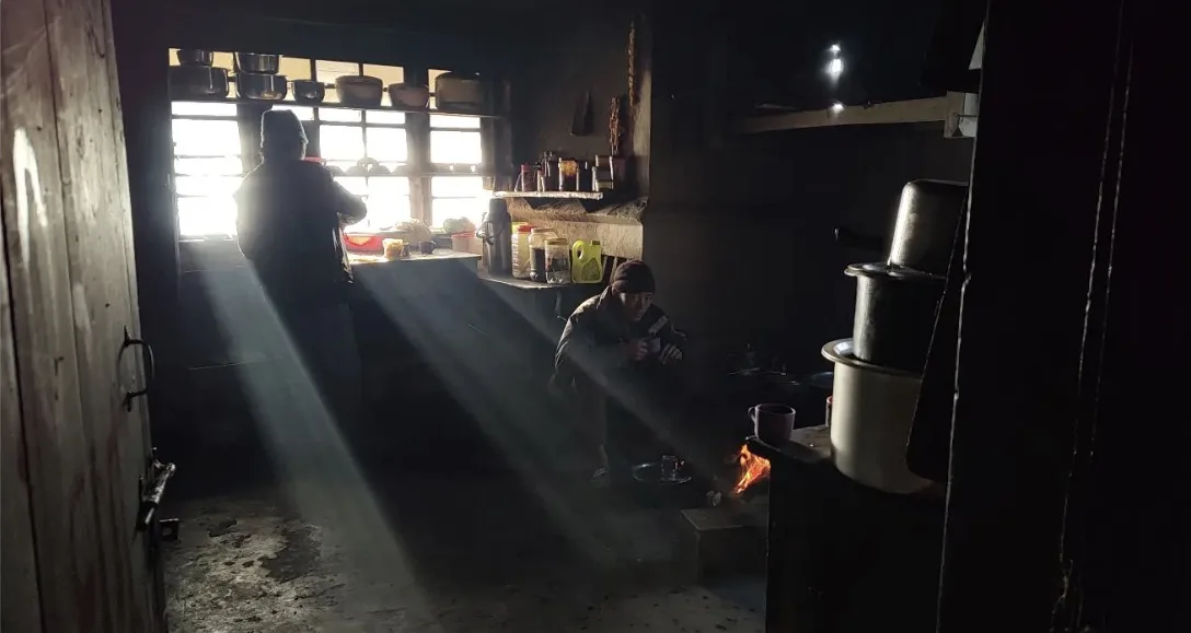 A dark mountain kitchen with shafts of morning light falling across a man crouched by a fire