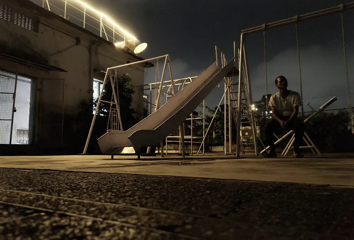 A rooftop playground at night, a slide and swings lit by a string of lights, one man sitting alone