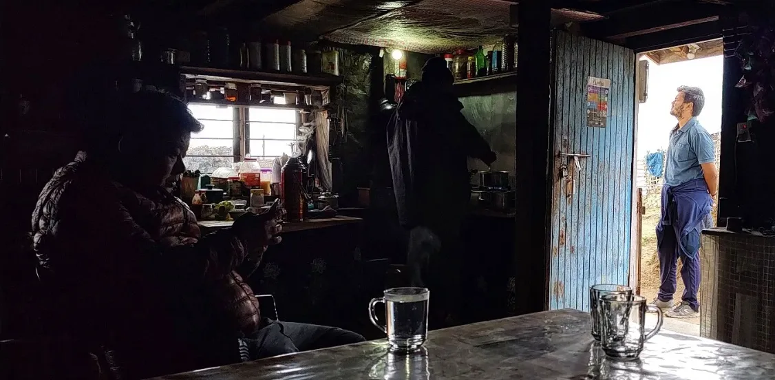 Inside a dim teahouse, glasses of tea on a table, a man in the bright doorway looking up at the sky