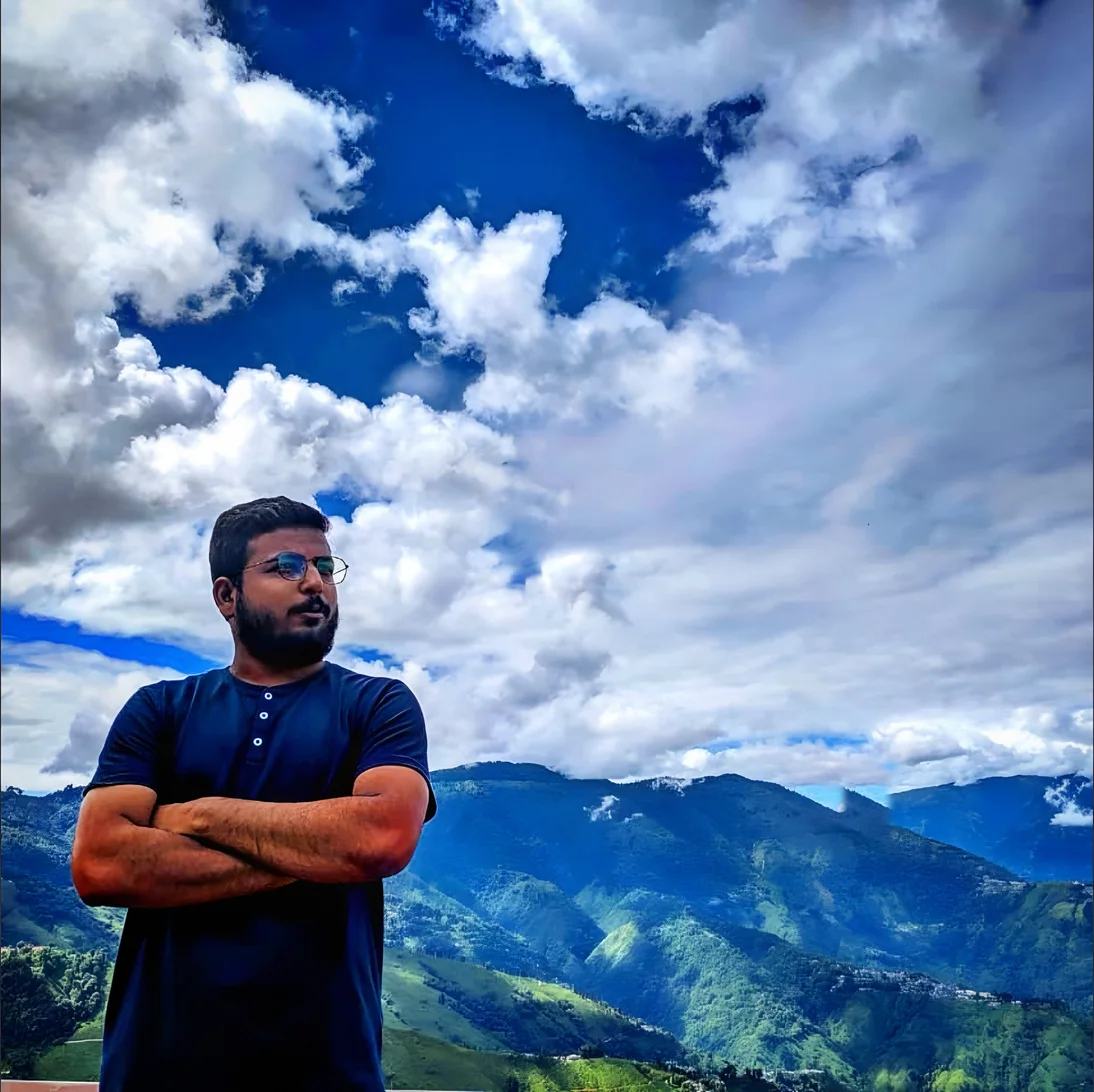 Sumedh with arms crossed under a huge sky of cloud above the green hills of Darjeeling